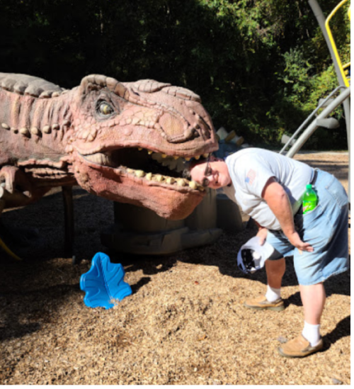 A person playfully bending down near a large dinosaur sculpture in a playground setting.