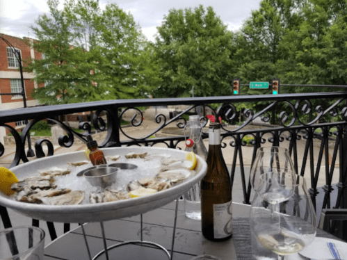 A seafood platter with oysters on ice, lemon wedges, and wine glasses, set on a balcony overlooking a street.