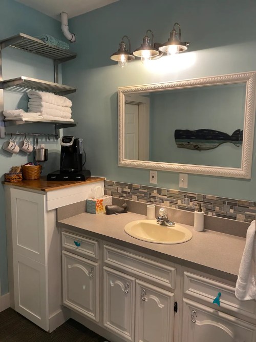 Bright bathroom with a light blue wall, white cabinetry, a mirror, and neatly arranged towels and coffee maker.