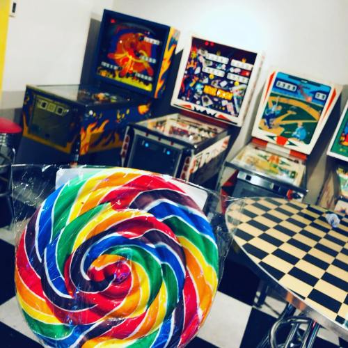 A colorful lollipop in the foreground with vintage pinball machines and a checkered table in the background.