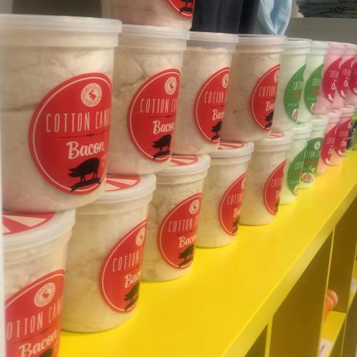 Containers of cotton candy labeled "Bacon" in various colors, arranged on a yellow shelf.