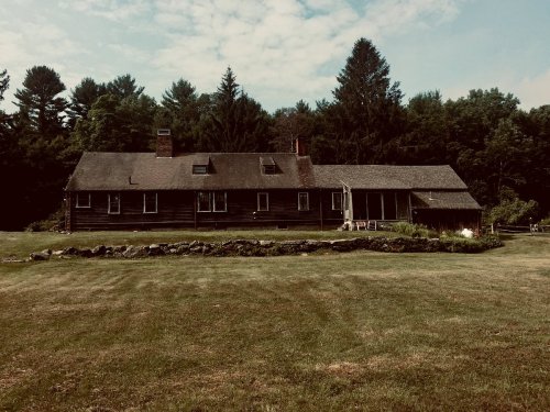A rustic wooden house surrounded by a grassy lawn and trees under a cloudy sky.