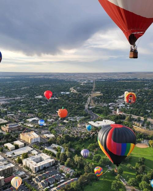 Aerial view of colorful hot air balloons over a green landscape and city, with a cloudy sky in the background.