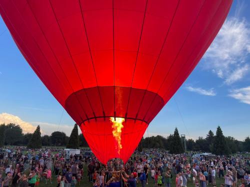 A vibrant red hot air balloon inflates above a large crowd at an outdoor event, with flames visible from the burner.