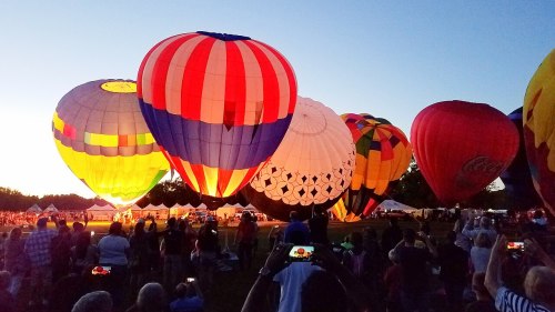 Colorful hot air balloons illuminated at dusk, with a crowd watching in the foreground.