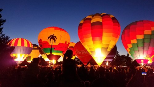 Colorful hot air balloons illuminated at night, surrounded by a crowd, with a clear sky in the background.