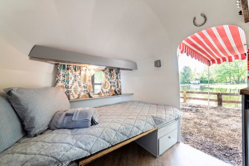 Cozy interior of a camper with a bed, colorful curtains, and a view of a lake through an arched doorway.