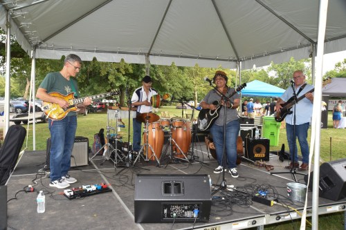 A band performs on stage under a tent, featuring guitarists, a drummer, and a vocalist, with an audience in the background.