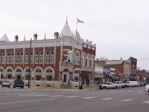 Historic brick building with a decorative roof, located at a street corner in a small town, with parked cars nearby.