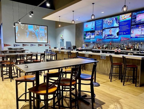 A modern bar with high tables, chairs, and multiple screens showing sports events. Bright and inviting atmosphere.