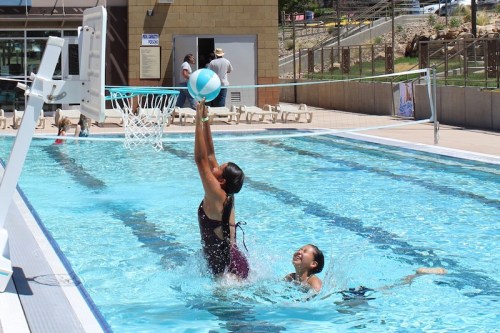 A girl jumps to make a basket in a pool while another girl swims nearby, both enjoying a sunny day.