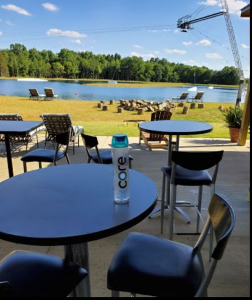 A serene lakeside view with empty tables and chairs, featuring a water bottle on a table in the foreground.