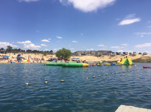A sunny beach scene with people enjoying water activities, inflatable structures, and a sandy shore in the background.