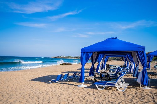 Blue beach cabanas and lounge chairs on sandy shore with gentle waves and clear blue sky.