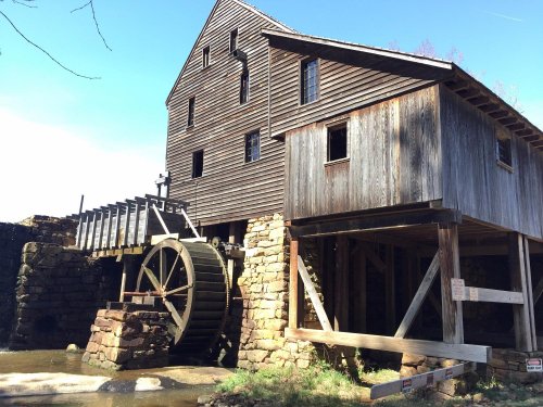 Historic wooden mill with a large water wheel, set against a clear blue sky and surrounded by greenery.
