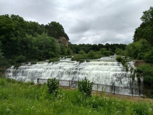 A cascading waterfall surrounded by lush greenery and trees under a cloudy sky. A fence lines the foreground.