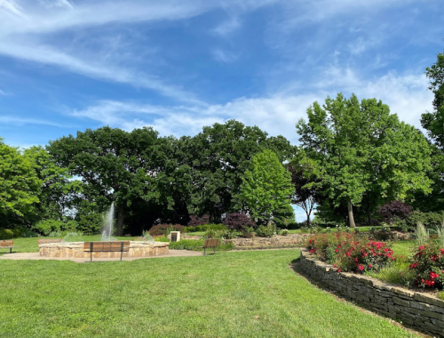 A serene park scene featuring a fountain, lush green grass, trees, and flower beds under a blue sky.
