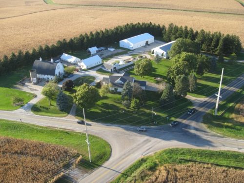 Aerial view of a rural farmstead with multiple buildings, surrounded by fields and intersecting roads.