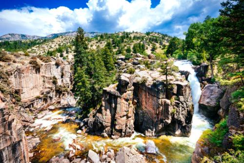 A scenic waterfall cascades over rocky cliffs, surrounded by lush greenery and mountains under a bright blue sky.