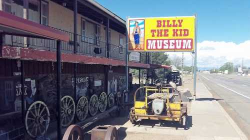 Sign for Billy the Kid Museum with vintage wagons and a yellow cart outside a building along a road.