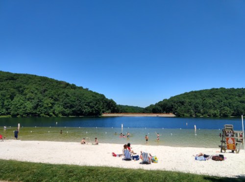 A sandy beach by a calm lake surrounded by green hills, with people enjoying the sun and swimming. Clear blue sky above.