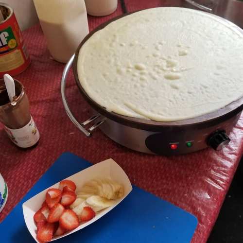A crepe maker with a smooth batter, alongside a plate of sliced strawberries and bananas on a blue cutting board.