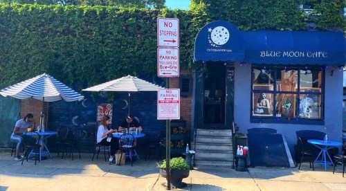 A cozy café with outdoor seating, blue awnings, and a mural, surrounded by parking signs and greenery.