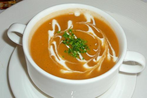 A white cup filled with creamy orange soup, garnished with a swirl of cream and fresh green herbs on top.