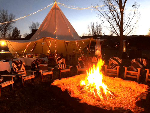 A cozy outdoor setting with a fire pit, surrounded by chairs and a tent, illuminated by string lights at dusk.