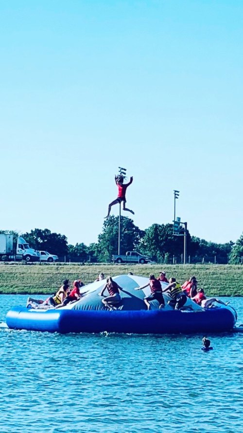 A group of people on a blue inflatable water toy, with one person jumping high above them against a clear blue sky.