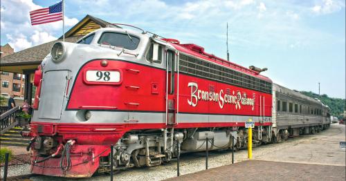 A vintage red and silver train at a station, with an American flag in the background and a cloudy sky above.