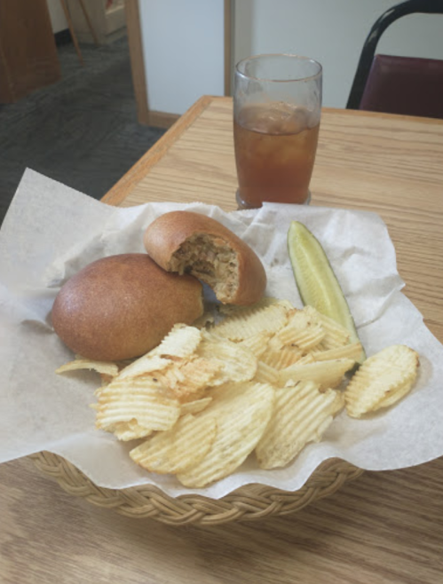 A basket with two bread rolls, a pickle, potato chips, and a glass of iced tea on a wooden table.