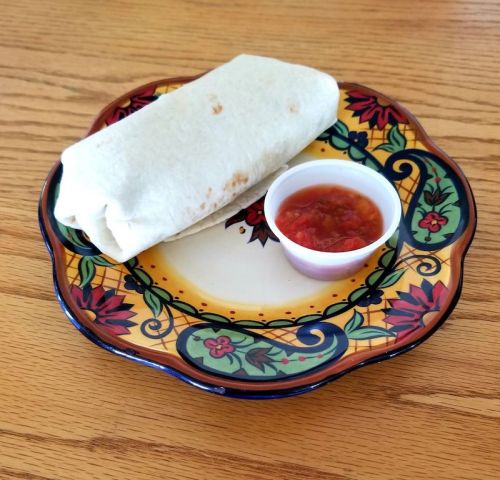 A flour tortilla burrito on a decorative plate, accompanied by a small cup of salsa.