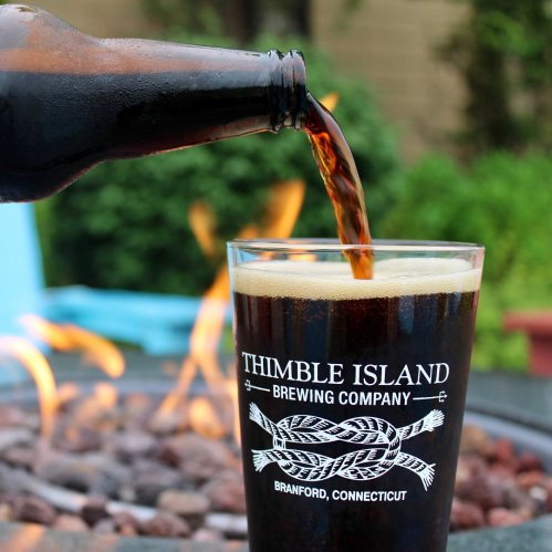 A bottle pours dark beer into a glass with "Thimble Island Brewing Company" label, set against a green background.
