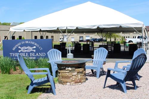 Outdoor seating area with blue chairs around a stone fire pit, under a large tent with a sign for Thimble Island Brewery.