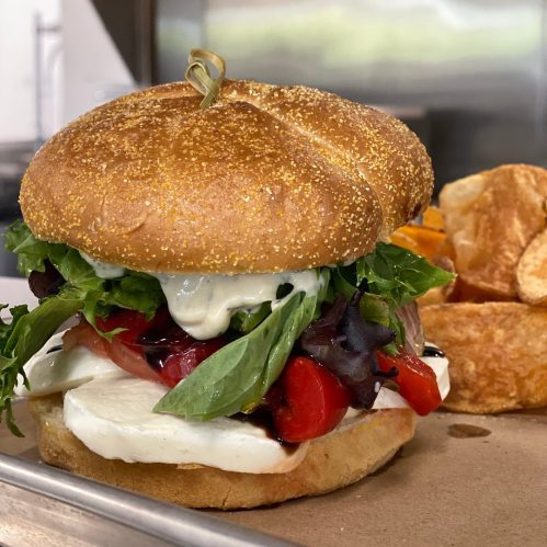 A close-up of a sandwich with a sesame seed bun, fresh vegetables, mozzarella, and creamy dressing.