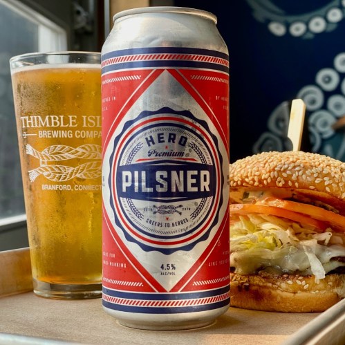 A can of Pilsner beer next to a glass of beer and a sesame seed bun burger with lettuce and tomato.