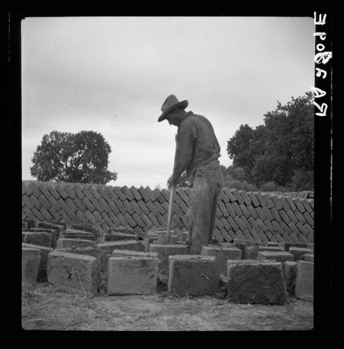 A man in a hat works with a tool among stacked bricks in a rural setting, with trees in the background.
