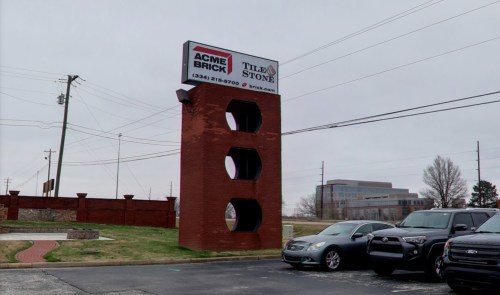 Sign for Acme Brick and Tile & Stone on a brick structure, with parked cars and a cloudy sky in the background.