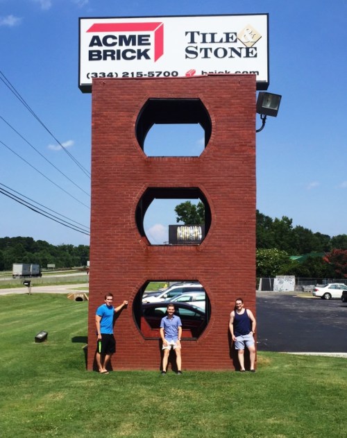 Three people pose in front of a large brick structure with three circular cutouts, under a sign for ACME Brick.