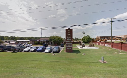 A brick building with a sign, surrounded by parked cars and green grass, under a clear sky. Power lines in the background.