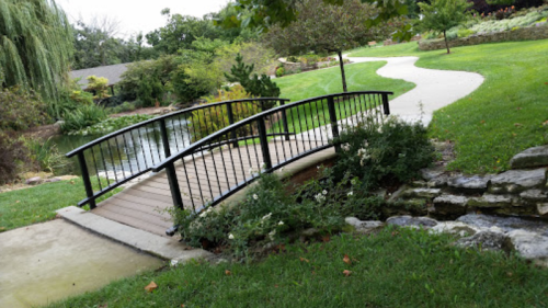 A small wooden bridge over a pond, surrounded by lush greenery and a winding path in a serene park setting.