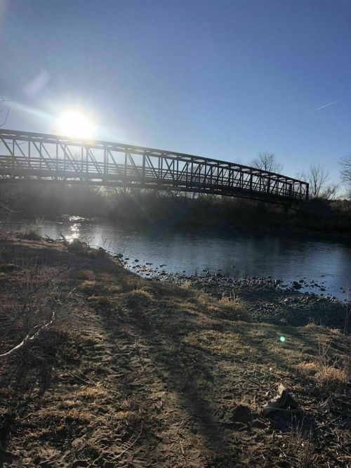 A sunlit bridge spans a river, with a sandy bank and trees in the background under a clear blue sky.