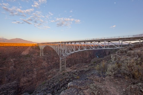 A large metal bridge spans a deep canyon at sunset, with rocky terrain and a clear sky in the background.