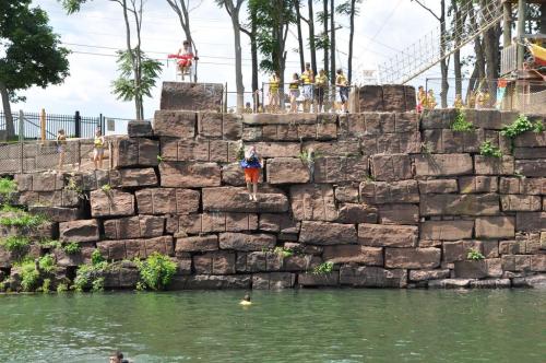 A person prepares to jump off a rocky ledge into a lake, with onlookers and trees in the background.