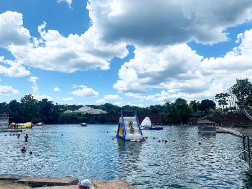 A sunny day at a lake with people swimming, inflatable structures, and fluffy clouds in the blue sky.
