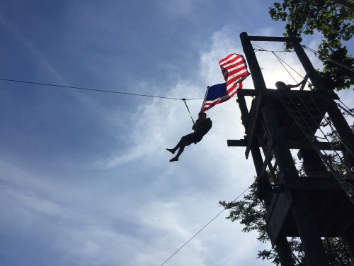 A person zip-lining while holding an American flag against a blue sky with wispy clouds.