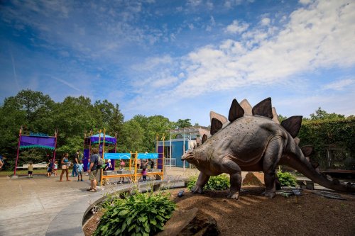 A large dinosaur statue in a park with playground equipment and people enjoying the outdoor space under a blue sky.