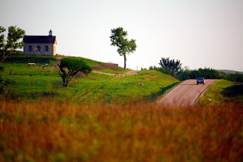 A scenic view of a rural landscape featuring a small house, a tree, and a winding road through green fields.