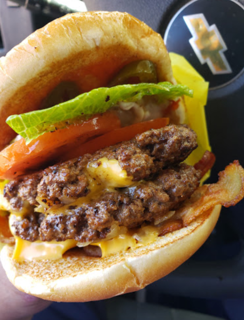 Close-up of a juicy burger with two beef patties, cheese, lettuce, tomato, and pickles, held in a hand.